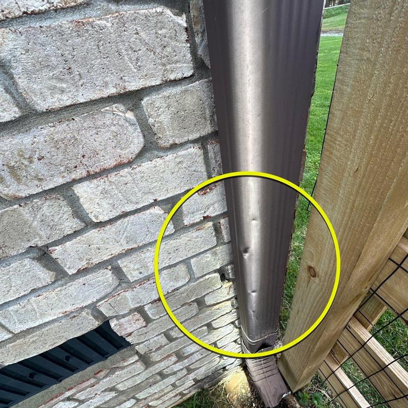 Metal downspout dent damage on exterior wall near fence
