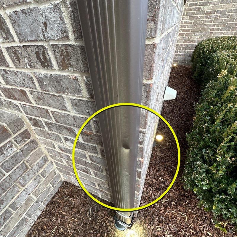 Downspout with dent damage near brick wall and shrubbery