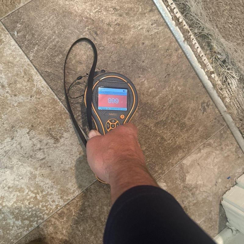 Moisture meter measuring tile floor dampness near baseboard