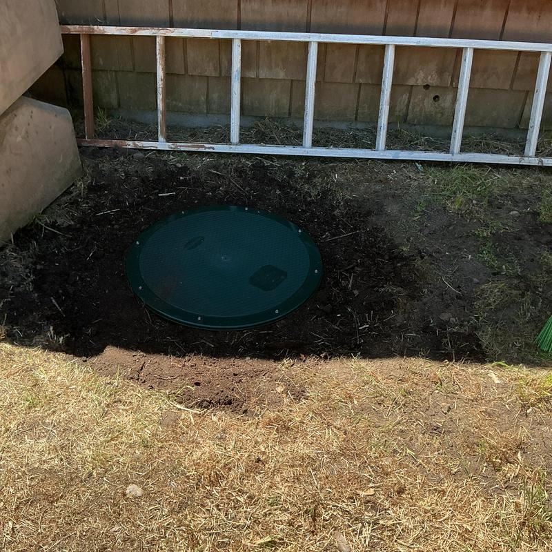 Septic tank cover in cleared soil beside house and ladder