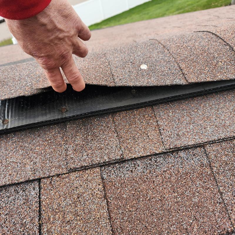 Asphalt roof shingles lifted revealing damaged ridge cap underlayment