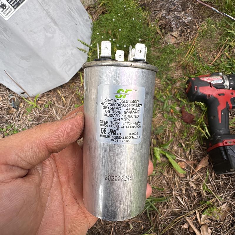AC capacitor held in hand with drill on grass nearby